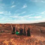 Group of five adventurers raise arms atop striking red rocks beneath vibrant blue sky on 7 Day Adelaide to Uluru Outback Tour.