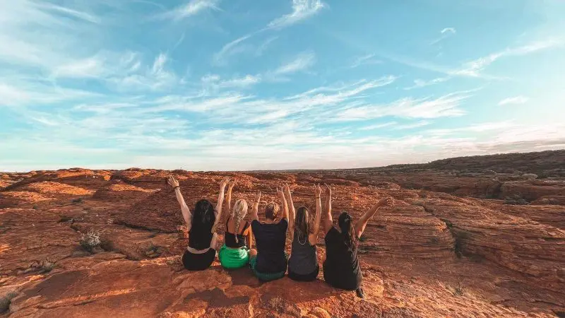 Group of five adventurers raise arms atop striking red rocks beneath vibrant blue sky on 7 Day Adelaide to Uluru Outback Tour.