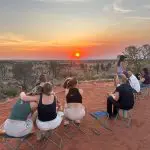 Travellers on the 7 Day Adelaide to Uluru Adventure by Untamed Escapes watch a stunning sunset over vibrant red desert scenery.