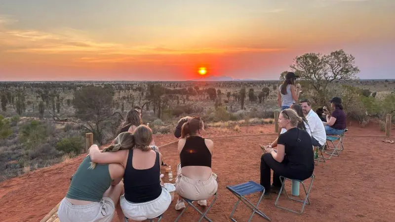 Travellers on the 7 Day Adelaide to Uluru Adventure by Untamed Escapes watch a stunning sunset over vibrant red desert scenery.