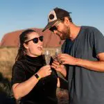 Two travellers wearing sunglasses toast champagne at iconic rock formations during a 7-Day Adelaide to Uluru Adventure Untamed Escapes.