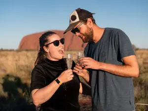 Two travellers wearing sunglasses toast champagne at iconic rock formations during a 7-Day Adelaide to Uluru Adventure Untamed Escapes.
