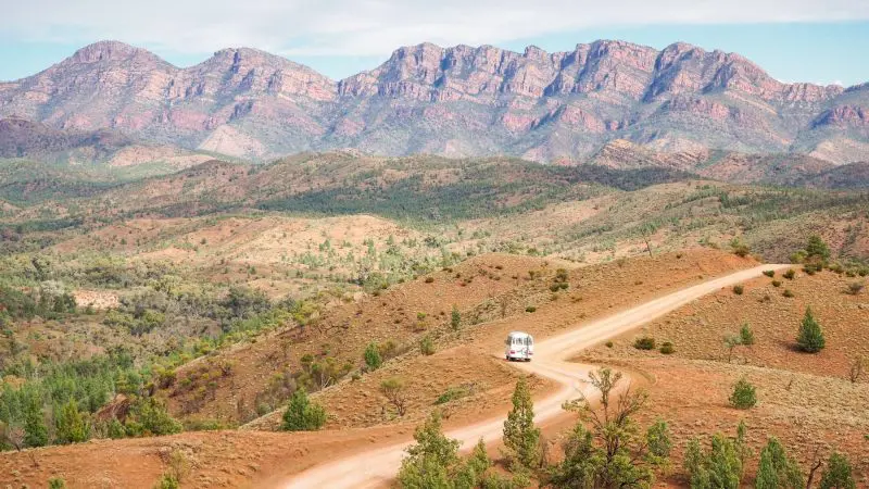 White campervan travels scenic hills on a 7-day Adelaide to Uluru adventure, Untamed Escapes tour, with distant mountain views.