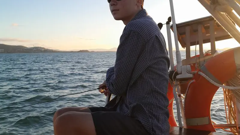 A young person wearing sunglasses and a cap sails on a Townsville Lunchtime Private Charter, enjoying a vibrant sunset on the boat.