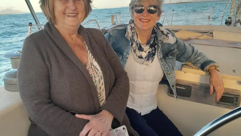 Two women happily smile aboard a sailboat on a Townsville Lunchtime Sailing Private Charter, with blue ocean waves and clear sky views.