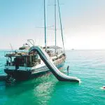 Luxury yacht with water slide into vibrant turquoise sea, people paddleboarding and swimming nearby beneath a sunny blue sky.