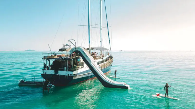 Luxury yacht with water slide into vibrant turquoise sea, people paddleboarding and swimming nearby beneath a sunny blue sky.