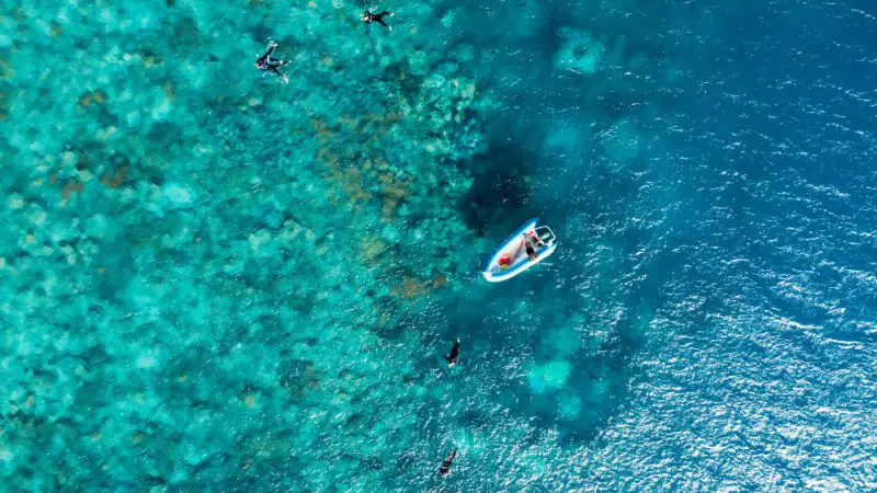 High-resolution aerial shot of snorkellers by a small boat above crystal-clear blue-green water and vibrant coral reef.