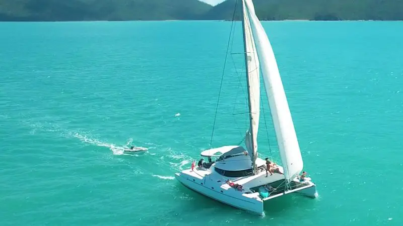 Luxury white catamaran glides on turquoise Whitsunday waters near lush green hills—Entice 2 Day 2 Night Sailing adventure in sun.