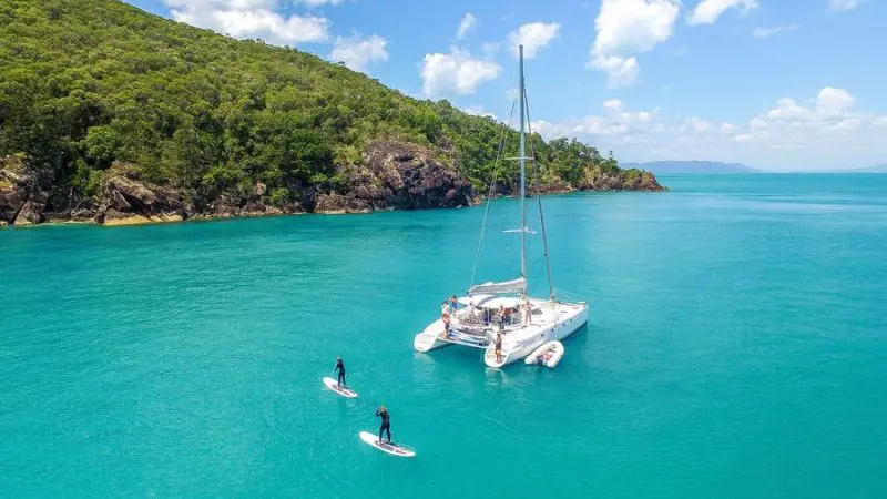 Whitsunday catamaran anchored in crystal-clear turquoise water, paddleboarders enjoying sunshine and blue skies, perfect sailing holiday.