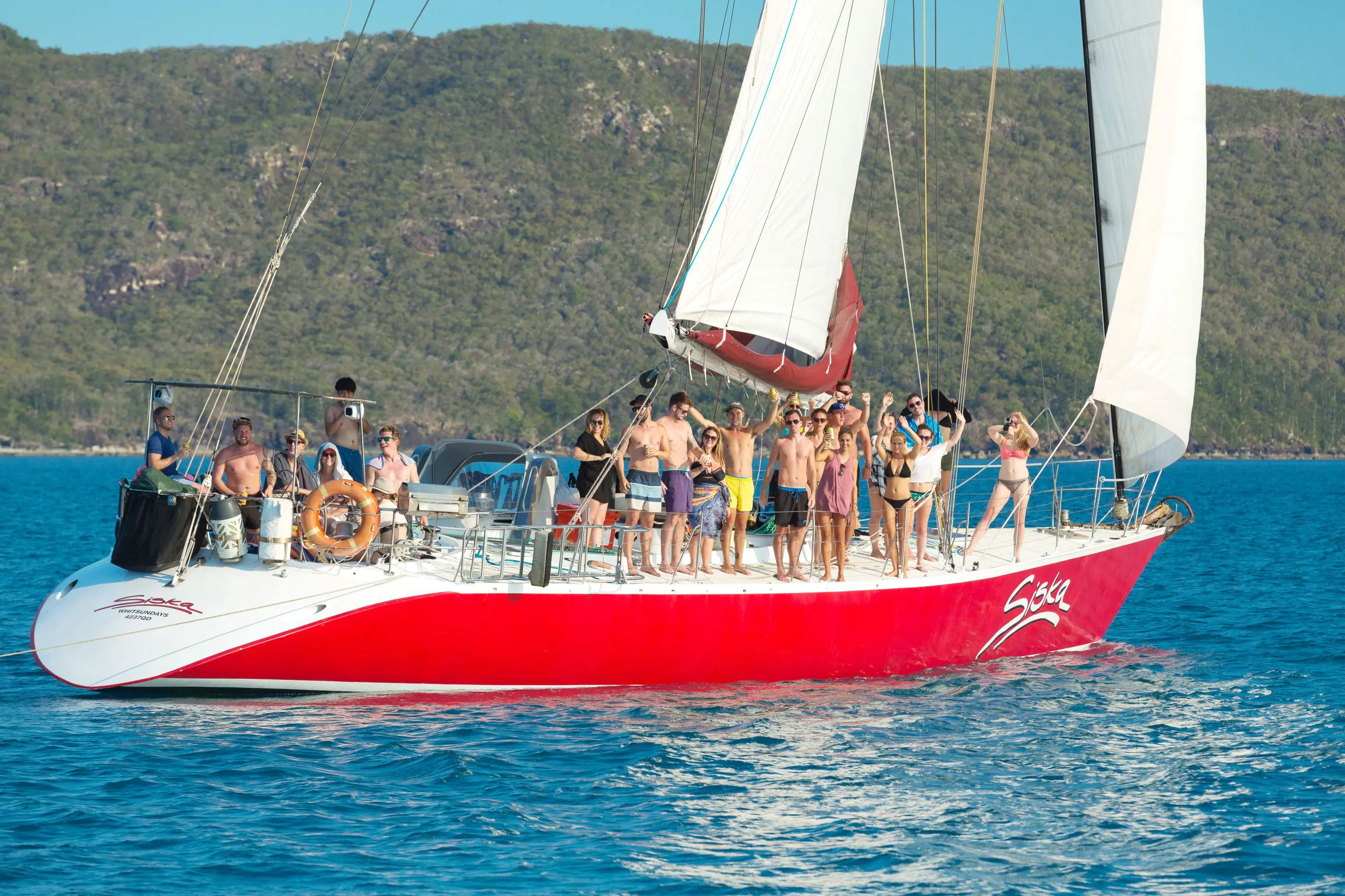 Vibrant group in swimwear waves from a striking red sailboat on the Siska 2 Day 1 Night Whitsundays Sailing adventure tour.