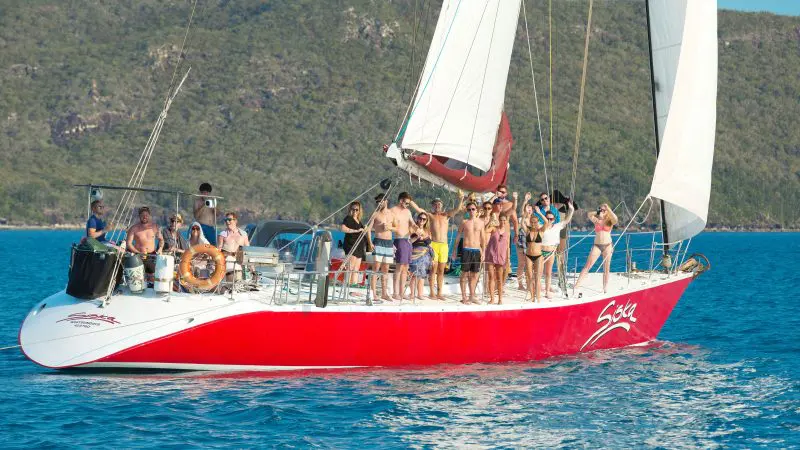 Vibrant group in swimwear waves from a striking red sailboat on the Siska 2 Day 1 Night Whitsundays Sailing adventure tour.