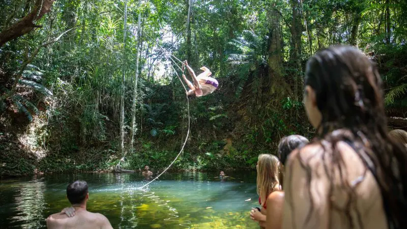 Adventurer swings on a rope over a lush forest pond during the renowned 1 Day Cape Tribbbin Tour With Uncle Brian in Queensland.