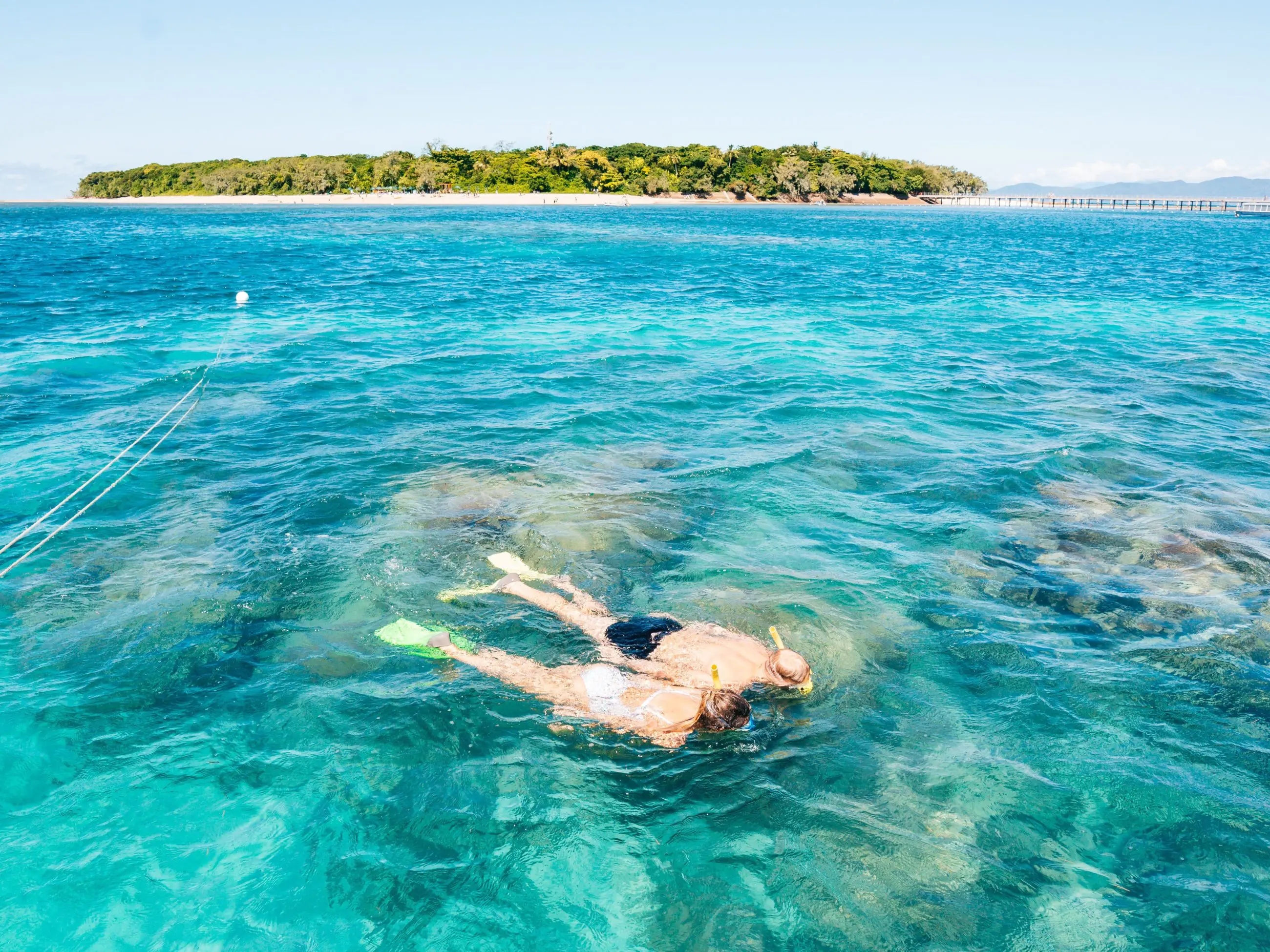 Two snorkellers and a glass-bottomed boat at Green Island above vibrant coral reef, gliding over crystal-clear blue waters in Cairns, Australia.