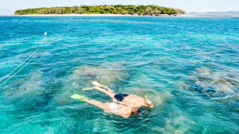 Two snorkellers and a glass-bottomed boat at Green Island above vibrant coral reef, gliding over crystal-clear blue waters in Cairns, Australia.