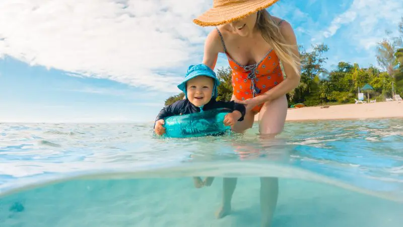Smiling woman in straw hat assists toddler paddling in crystal-clear Green Island waters, sunny beach backdrop, perfect family day.