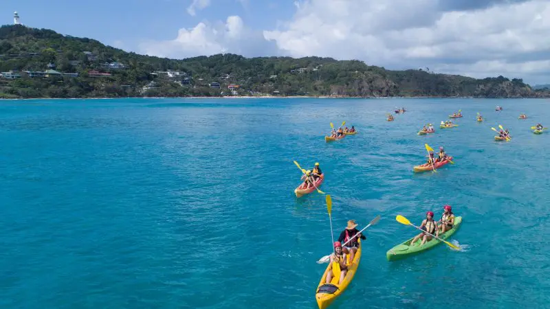 Adventurers kayaking in crystal-clear blue sea waters on a guided tour, passing stunning coastal hills and picturesque seaside houses.