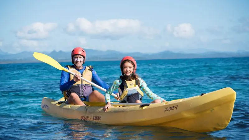 Two adventurers in helmets and life jackets paddle a yellow kayak on a guided sea kayaking tour across crystal clear blue waters.