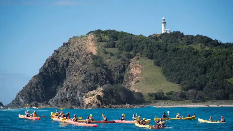 Guided sea kayaking tour group paddling by rugged green cliffs and a picturesque lighthouse under clear blue skies, perfect for adventure.