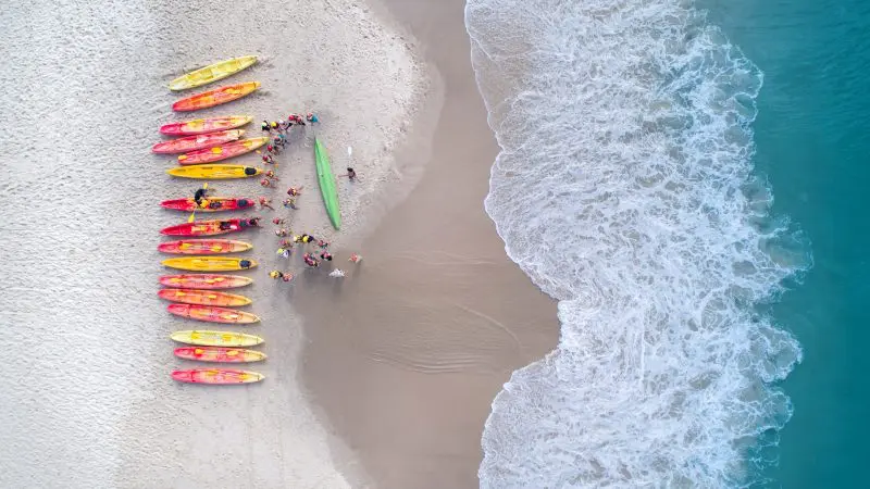 Aerial view of guided ocean kayaking tour, kayaks lined on sandy beach, group prepares for dolphin kayaking adventure, crystal blue water.