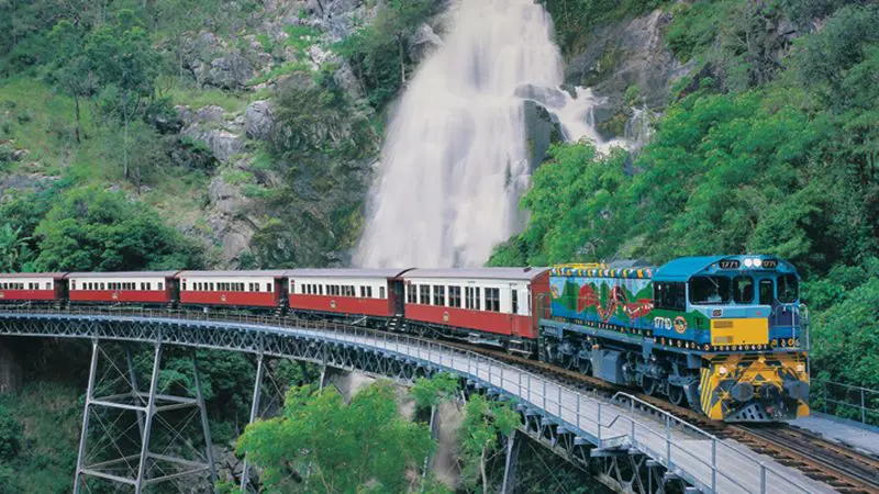 Kuranda Scenic Railway’s colourful Kuranda Deluxe train crosses a bridge near a waterfall and vibrant, lush green rocky hills in Australia.