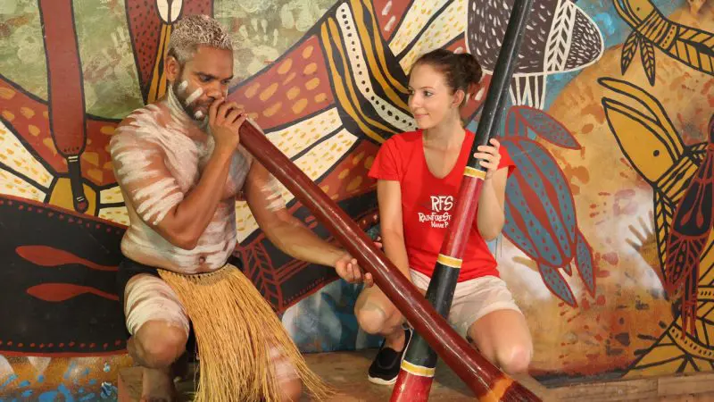 Indigenous Australian man playing didgeridoo as woman in red shirt observes, featured during Express Kuranda Later Departure tour.