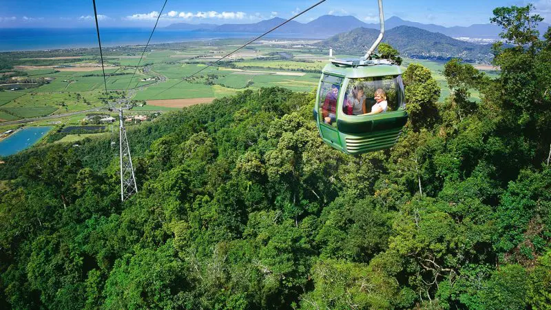 Experience breathtaking panoramic views as a green cable car glides over the lush Kuranda forest, ocean and blue sky beyond.