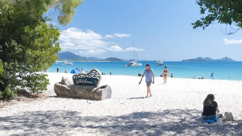 Visitors unwind on Whitehaven Beach’s pristine white sand beside crystal-clear turquoise waters, with tour boats anchored offshore.