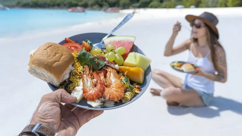 Gourmet seafood, fresh fruit, and artisan bread on a plate at Whitehaven Beach; woman enjoys meal by the shore after snorkelling.