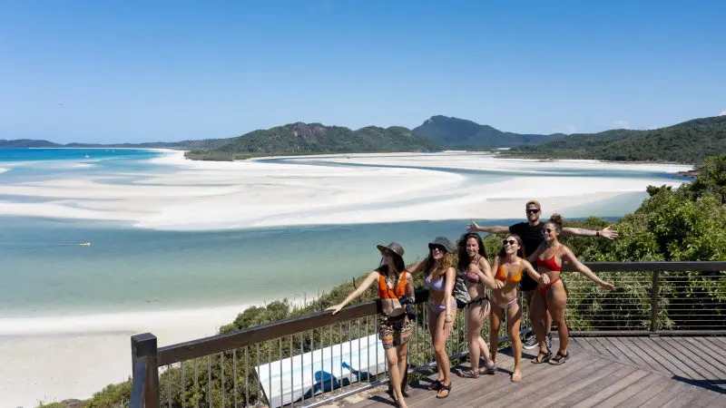 Six friends in colourful swimsuits smile on a sunlit wooden deck, enjoying a Big Fury Whitsundays tour at Whitehaven Beach, Australia.