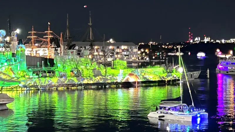 Sydney Harbour illuminated at night with boats and a vibrant Vivid Lights Catamaran Cruise glowing green and purple on the water.