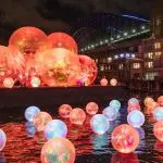Glowing light orbs reflect on Sydney Harbour’s waters at night during a Vivid Lights Catamaran Cruise, Sydney skyline in background.