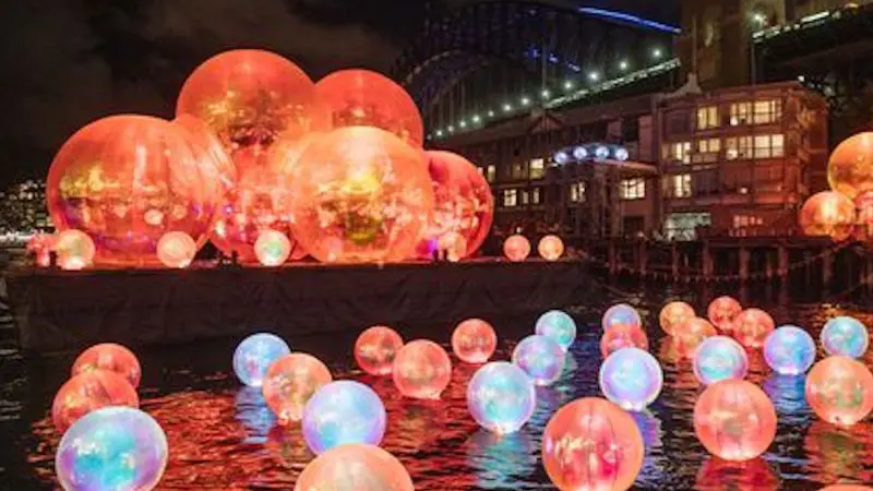 Glowing light orbs reflect on Sydney Harbour’s waters at night during a Vivid Lights Catamaran Cruise, Sydney skyline in background.