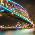 Sydney Harbour Bridge illuminated by Vivid Sydney lights at night, with a BYO catamaran cruise and the Opera House in the background.