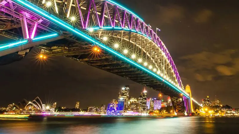 Sydney Harbour Bridge illuminated by Vivid Sydney lights at night, with a BYO catamaran cruise and the Opera House in the background.