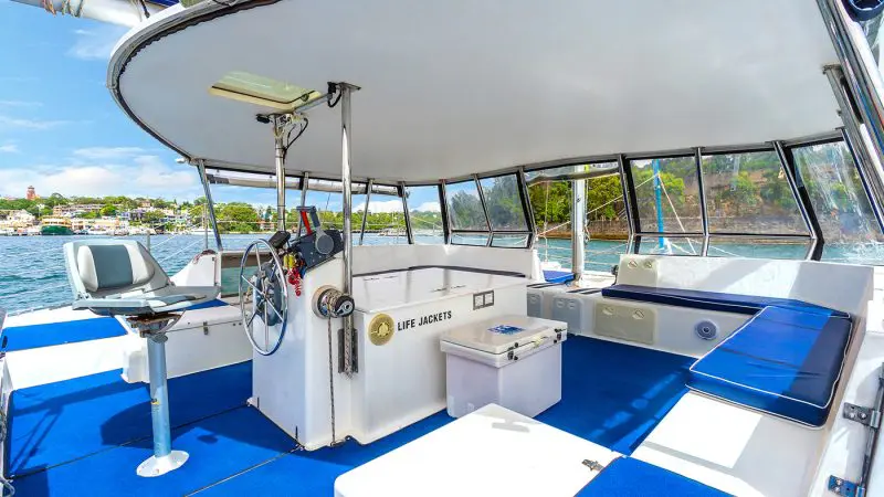 Catamaran cruise interior with blue seats, steering wheel, life jacket box, and panoramic views of iconic Sydney Harbour landmarks.