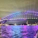Sydney Harbour Bridge illuminated at night, vibrant lights mirrored on water, viewed from a luxury cruise with gourmet canapés.