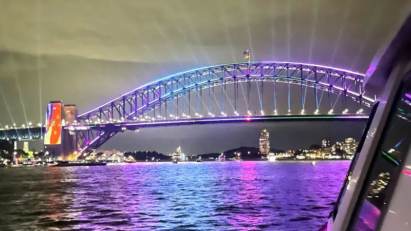 Sydney Harbour Bridge illuminated at night, vibrant lights mirrored on water, viewed from a luxury cruise with gourmet canapés.