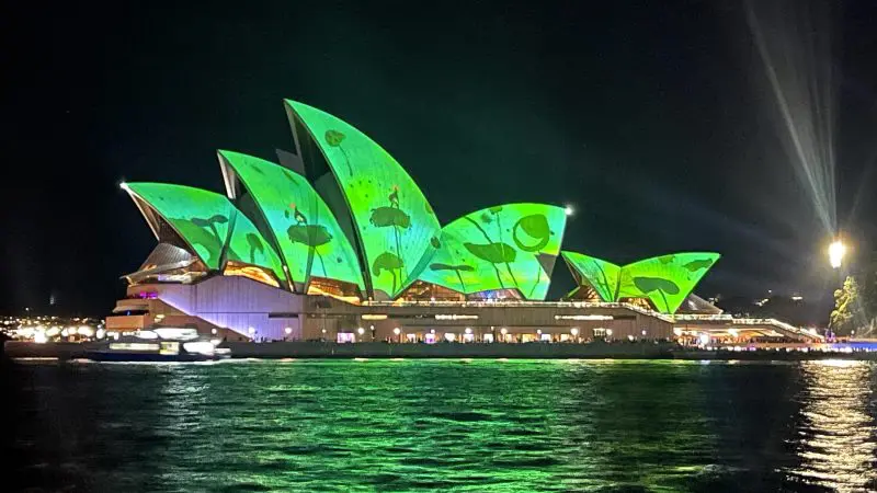 Sydney Opera House illuminated at night, viewed from a Sydney Harbour Vivid Lights cruise with canapés, reflecting vibrant green lights.