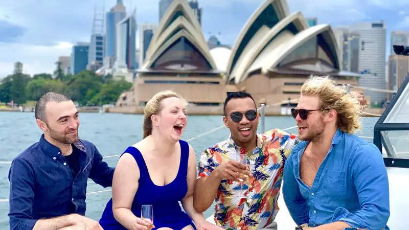 Group of friends laughing aboard a catamaran cruise with iconic Sydney Opera House and harbour landmarks in the scenic background.