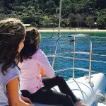 Two young girls enjoy a scenic catamaran cruise, admiring iconic Sydney Harbour views and a tree-lined beach under a clear sky.