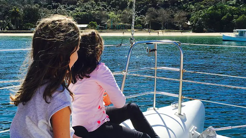 Two young girls enjoy a scenic catamaran cruise, admiring iconic Sydney Harbour views and a tree-lined beach under a clear sky.