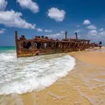 Discover the iconic rusty shipwreck on K'gari’s sandy beach, captured on a Noosa day tour beneath vibrant blue skies and wispy clouds.