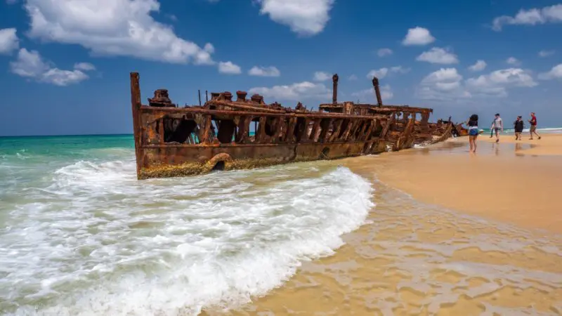 Discover the iconic rusty shipwreck on K'gari’s sandy beach, captured on a Noosa day tour beneath vibrant blue skies and wispy clouds.