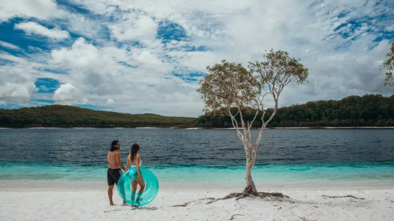 Two people holding a blue swim ring on a pristine sandy beach during K'gari Day Tour Ex Noosa, lush forest visible in the background.