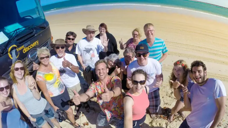 Tour group posing and smiling on Rainbow Beach during K'gari Day Tour with a black SUV, clear blue sky, bright sunny weather.
