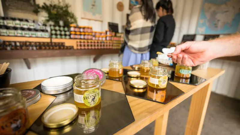 A guest samples fresh honey from jars on a rustic wooden table as Bruny Island Food Sightseeing visitors explore shelves behind.