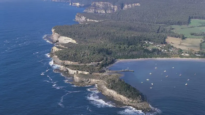 Stunning aerial view of Tasmania’s rugged coastline, lush forest, and tour boats on a Port Arthur Harbour Cruise and Lunch.