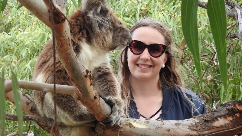 Smiling woman in sunglasses poses with a koala during a 6-Day Eyre Peninsula Flinders Ranges Adventure Tour in scenic outdoors.