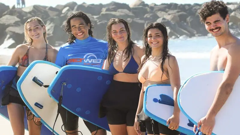 Five teenagers and young adults, aged 13 and over, smile with surfboards on a sunny beach before a two-hour beginner surfing lesson.